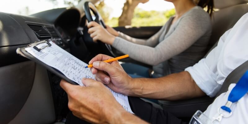 Examiner marking a driving test sheet while woman drives car